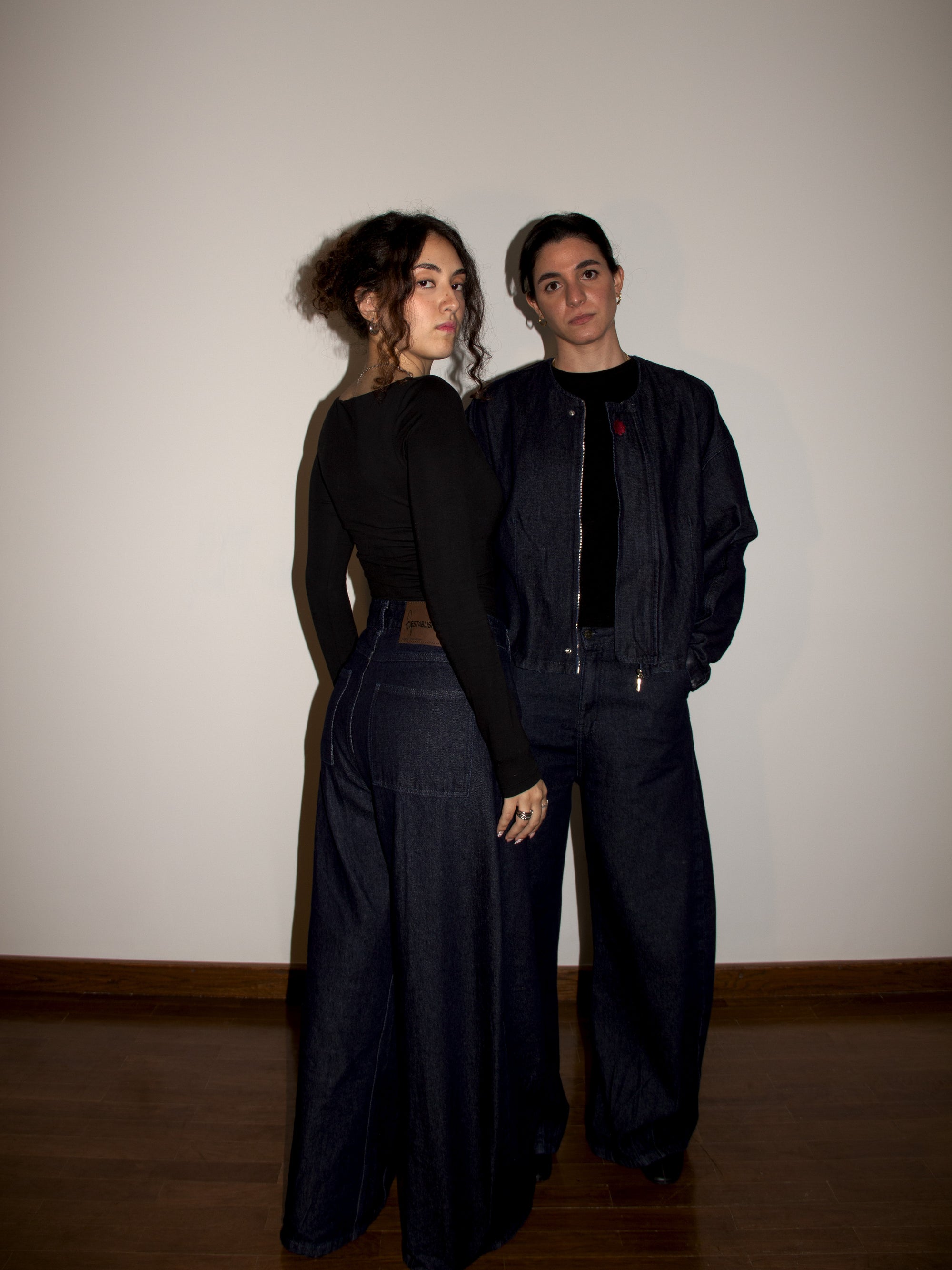 Two people wearing black outfits standing against a plain wall.