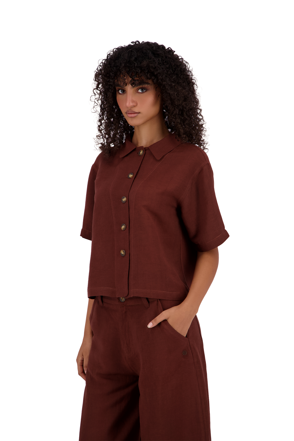 A model with curly dark hair wearing a matching rust-brown short-sleeve button-up shirt and wide-leg trousers, photographed against a solid white background. #Burnt_Clove