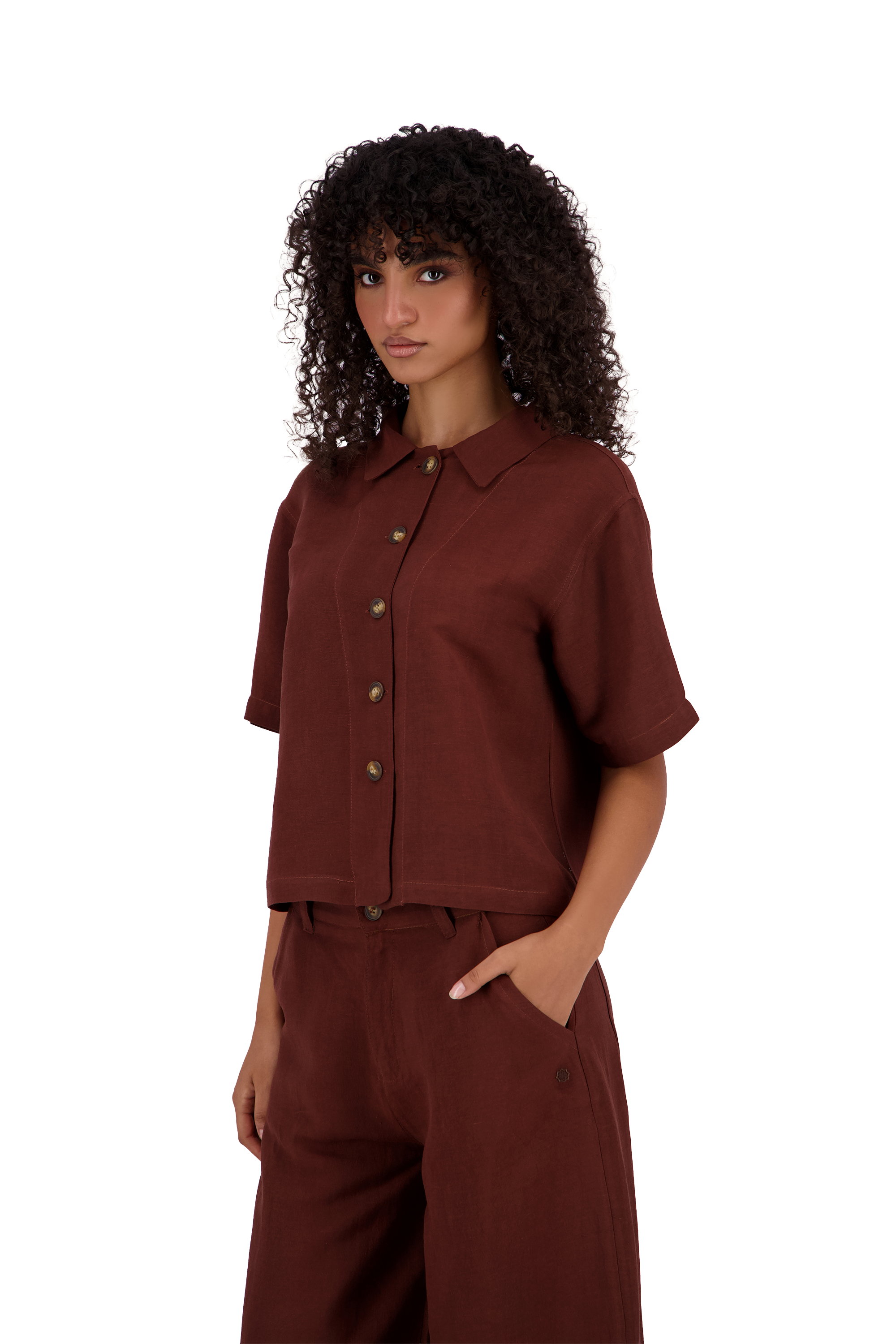 A model with curly dark hair wearing a matching rust-brown short-sleeve button-up shirt and wide-leg trousers, photographed against a solid white background. #Burnt_Clove