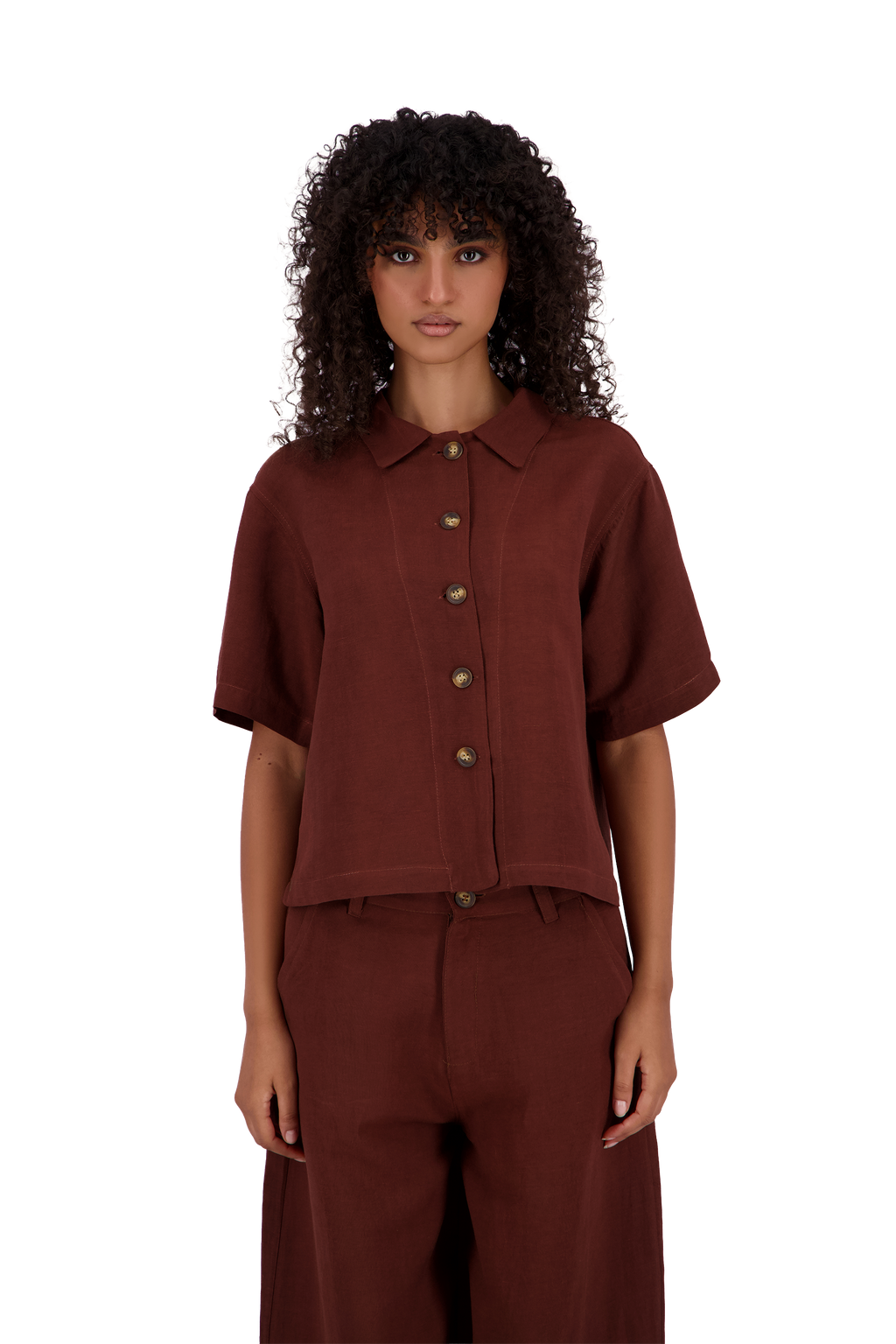 A model with curly dark hair wearing a short-sleeve button-front collared shirt and matching wide-leg trousers in a deep rust-brown color, shot against a plain white background. #Burnt_Clove