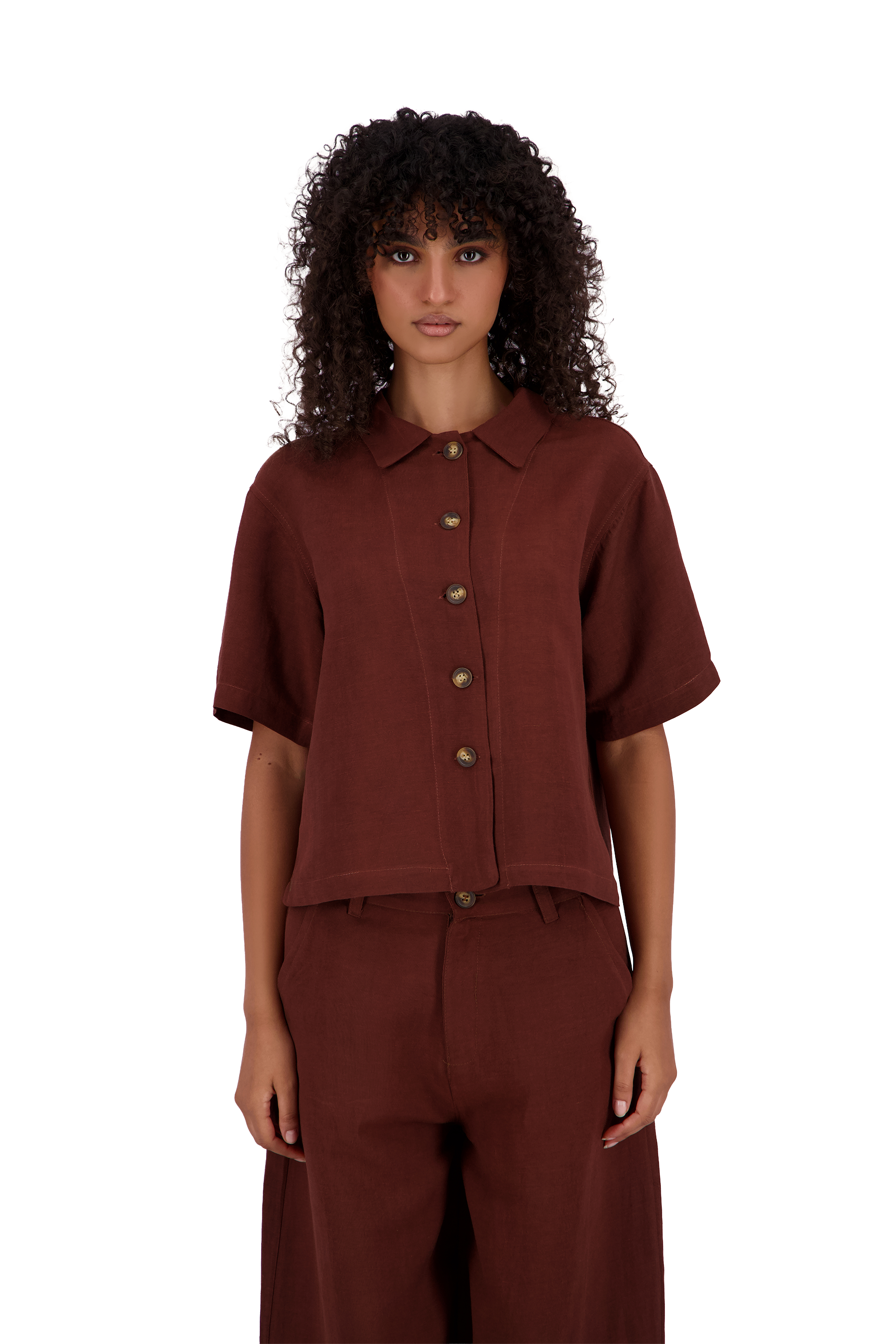 A model with curly dark hair wearing a short-sleeve button-front collared shirt and matching wide-leg trousers in a deep rust-brown color, shot against a plain white background. #Burnt_Clove