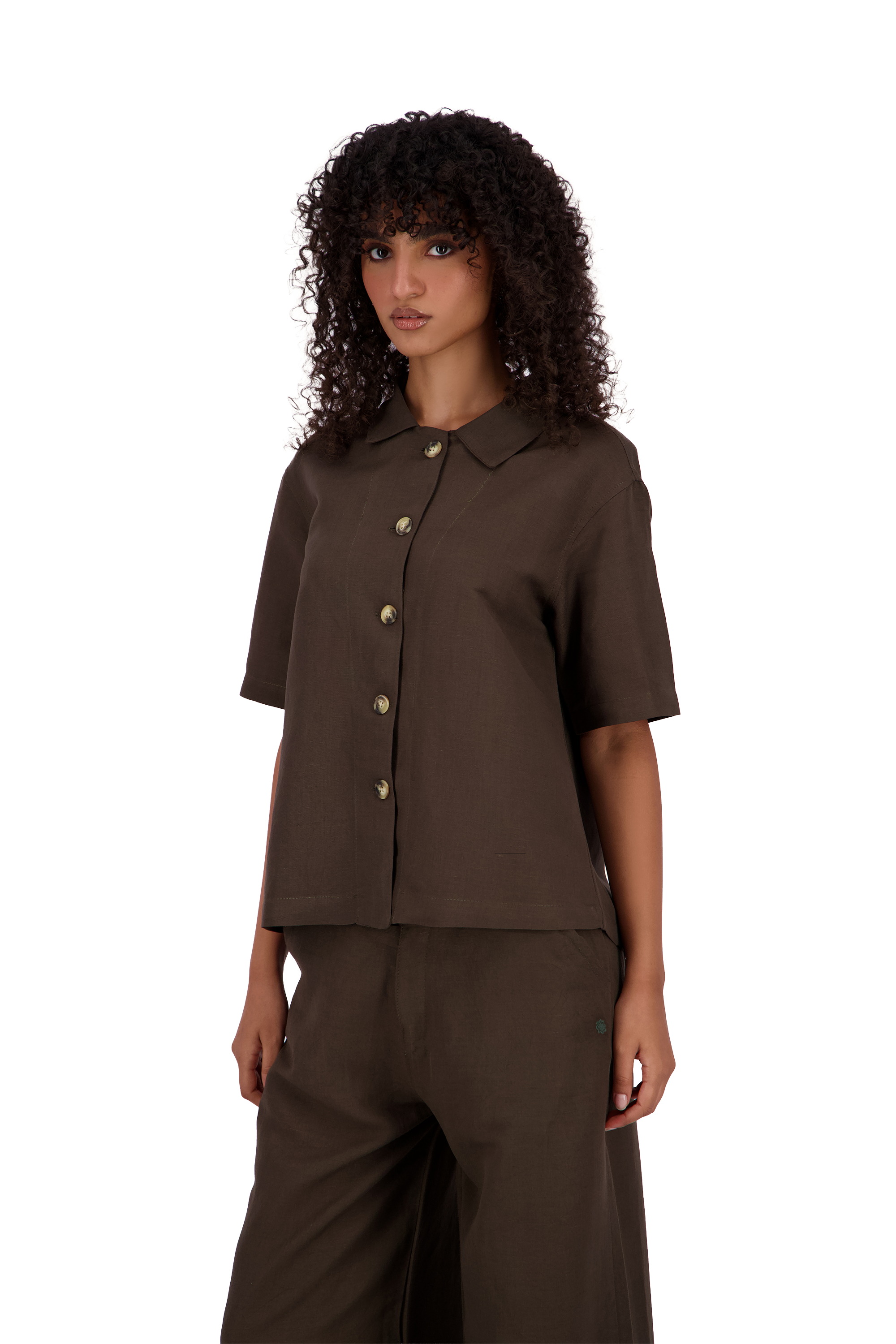 A model with curly dark hair wearing a dark brown short-sleeve button-up shirt and matching dark brown trousers, photographed against a solid white background. #Cumin_Leaf