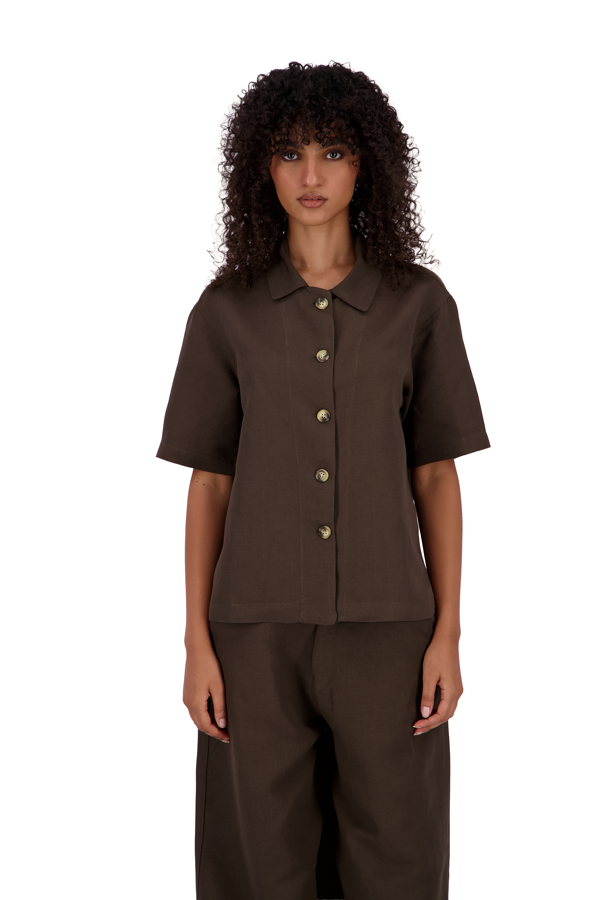 A model with curly dark hair wearing a dark brown short-sleeve button-up shirt and matching dark brown trousers, photographed against a solid white background. #Cumin_Leaf